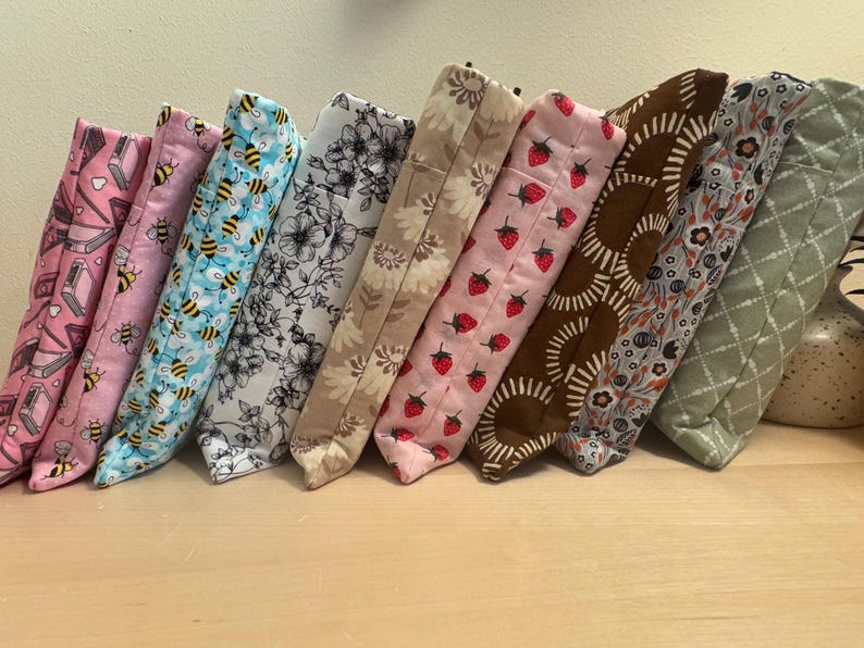 May include: Nine fabric pouches with different patterns, including bees, flowers, strawberries, and geometric designs. The pouches are lined up in a row on a light wood surface.