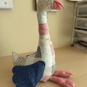 May include: A handmade goose-shaped stuffed animal. The goose features a patchwork design with floral and geometric patterns in pink, blue, and cream. It has a pink beak and feet, and blue wings. The goose is sitting on a wooden surface.