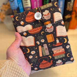 May include: A black book sleeve with a repeating pattern of teapots, books, cups, and flowers. The sleeve has a white button closure.