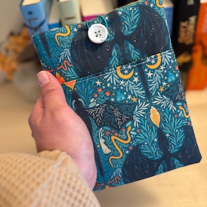 May include: A teal fabric book sleeve with a button closure. The sleeve features a dark blue bat, snake, rabbit, and celestial pattern. The button is white.