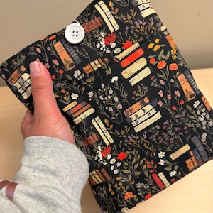 May include: A black fabric book sleeve featuring a floral and book pattern. The design includes stacks of books in various colors, along with flowers and foliage. A white button secures the sleeve closed.