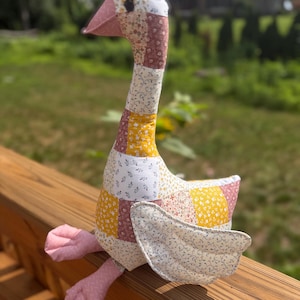 May include: A stuffed goose toy constructed from a patchwork of fabric squares featuring various floral and vine patterns in shades of yellow, pink, white, and brown. The goose has a solid pink beak and webbed feet with a dark circular eye.