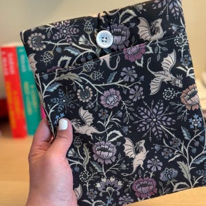 May include: A black fabric book sleeve with a floral and dragon pattern. The sleeve has a button closure and a pocket. The design includes flowers in shades of purple, pink, and blue, with beige dragons and foliage on a black background.