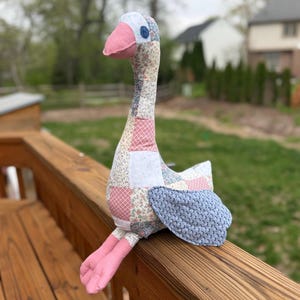 May include: A handmade goose plush toy with a patchwork design. The goose features a pink beak and feet, a blue eye, and a light blue wing. The body is made of fabric squares in various floral and geometric patterns.