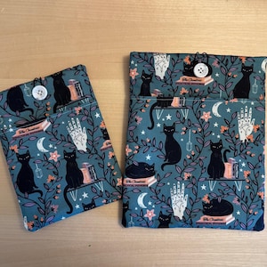 May include: Two teal blue fabric pouches with black cats, stars, and floral designs. The pouches have a button closure and the text "The Occult's Practical Potions" printed on them.
