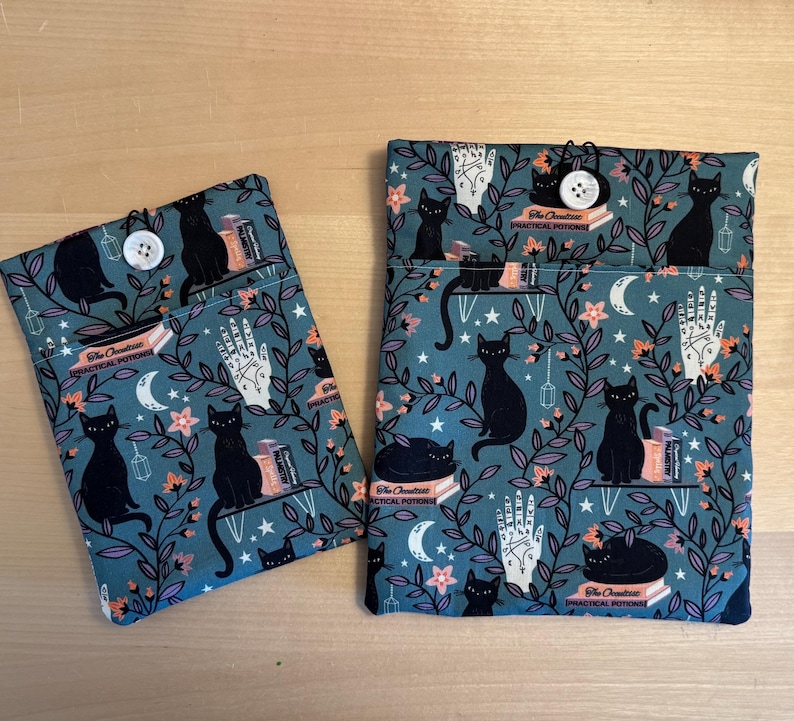 May include: Two teal blue fabric pouches with black cats, stars, and floral designs. The pouches have a button closure and the text "The Occult's Practical Potions" printed on them.