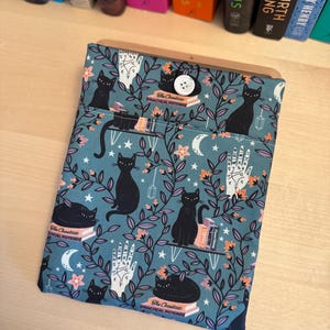 May include: A teal blue book sleeve with a black cat and hand print pattern. The sleeve has a pocket on the front and a button closure. The text "The Occultest Practical Potions" is printed on the sleeve.