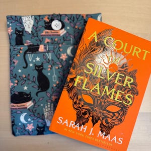 May include: A book cover for "A Court of Silver Flames" by Sarah J. Maas. The cover is orange with a black and gold mask design. The title is in gold lettering and the author's name is in white lettering.