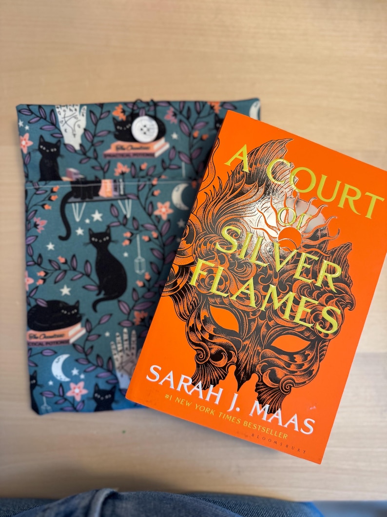 May include: A book cover for "A Court of Silver Flames" by Sarah J. Maas. The cover is orange with a black and gold mask design. The title is in gold lettering and the author's name is in white lettering.