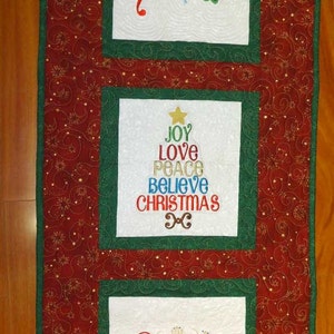 May include: A red, green, and white quilted table runner with embroidered Christmas words and a snowflake. The words include "Joy", "Believe", "Christmas", "Peace", and "Love".