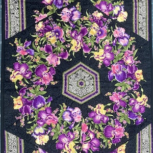May include: A black fabric with a repeating pattern of purple, pink, and yellow iris flowers. The fabric has a hexagonal design in the center with a white and green lace pattern.