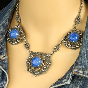 May include: A silver-toned necklace with three ornate, openwork pendants, each featuring a large, round, blue gemstone. The necklace is displayed on a mannequin wearing a denim jacket, showcasing the jewelry's design and color.
