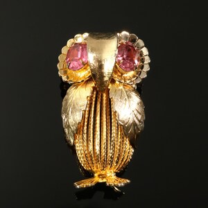May include: A gold-toned owl brooch with pink gemstone eyes. The brooch features detailed feather and leaf designs, with a textured body and a clasp on the back. The owl's head has two pink oval-shaped gemstones.