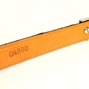 Puede incluir: Primer plano de una correa de cuero naranja claro con el n&uacute;mero "Q4800" estampado. La correa tiene un borde oscuro y un cierre de metal plateado con un acento rojo. El fondo es blanco.
