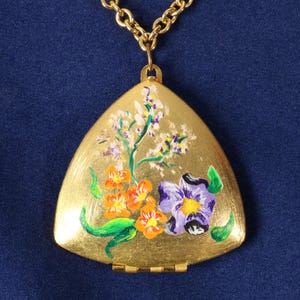 May include: A gold-toned triangular locket pendant suspended from a matching gold-toned chain. The front surface of the locket features a hand-painted floral design with orange, purple, and white flowers accented by green leaves on a brushed gold finish.