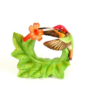 May include: A ceramic hummingbird figurine with a red head, green body, and brown wings. The bird is perched near an orange flower and green leaves. The figurine is designed to be a decorative accent.
