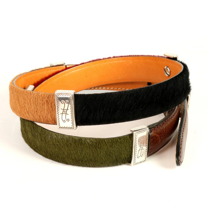 Puede incluir: Tres cinturones de cuero apilados con hebillas plateadas. El cintur&oacute;n superior tiene una textura de piel marr&oacute;n y negra, el del medio es negro y el inferior verde oliva. Las hebillas tienen un dise&ntilde;o grabado.