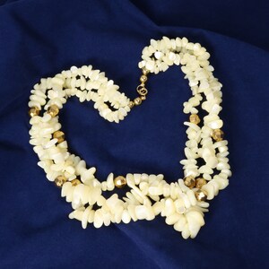 May include: A multi-strand necklace featuring numerous small, irregular, off-white shell chips interspersed with polished, faceted gold-tone metallic beads. The necklace is secured with a gold-tone metal clasp and circular connector beads.