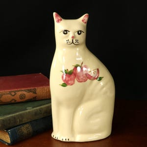 May include: Cream-colored ceramic cat figurine with pink accents on the ears and a painted face. The cat has a design of red apples with green leaves on its chest. The figurine is sitting upright.