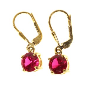 May include: A pair of gold-colored leverback earrings, each featuring a round, vibrant red gemstone. The gemstones are held in place by four prongs, adding a touch of elegance to the jewelry.