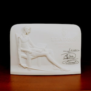 May include: A white porcelain relief sculpture featuring a seated figure holding a sword. The sculpture has the text "LLADRO COLLECTOR SOCIETY" and signatures. The figure is seated in a chair, with a book in their lap.