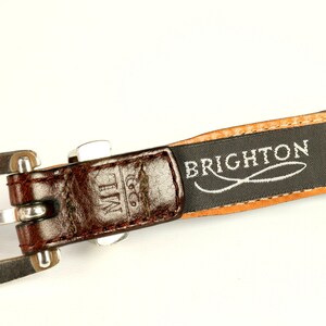 Puede incluir: Primer plano de un cintur&oacute;n de cuero marr&oacute;n con hebilla plateada. El cintur&oacute;n presenta el nombre de la marca "BRIGHTON" impreso en una etiqueta negra. Las iniciales "ML" est&aacute;n grabadas en el cuero.