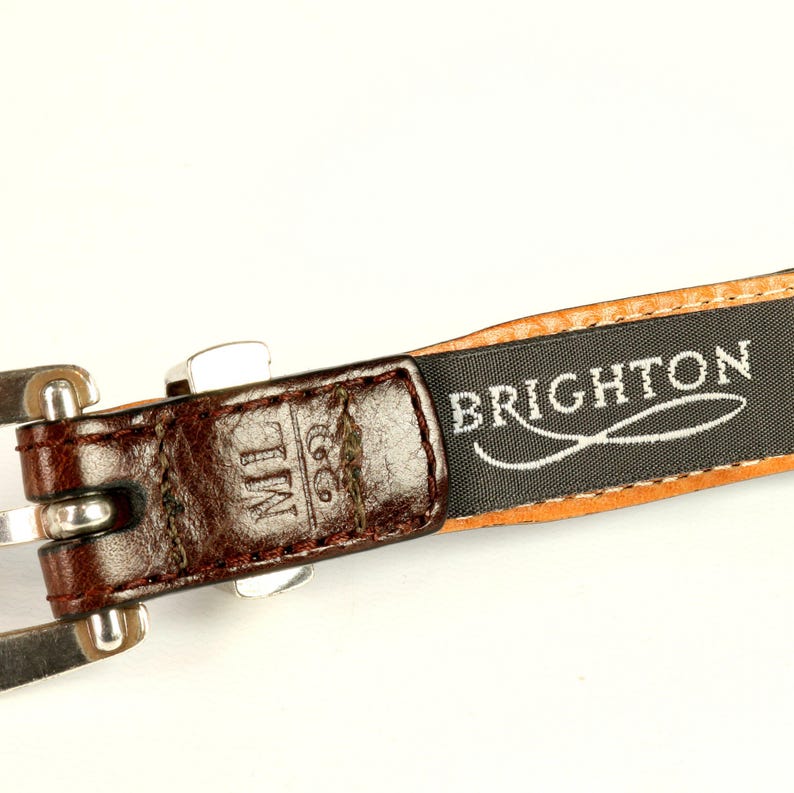 Puede incluir: Primer plano de un cintur&oacute;n de cuero marr&oacute;n con hebilla plateada. El cintur&oacute;n presenta el nombre de la marca "BRIGHTON" impreso en una etiqueta negra. Las iniciales "ML" est&aacute;n grabadas en el cuero.