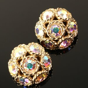 May include: A pair of gold-toned, round earrings with iridescent, multi-colored crystals. Each earring features a central crystal surrounded by smaller crystals and a twisted rope-like border. The crystals reflect hues of pink, yellow, and blue.