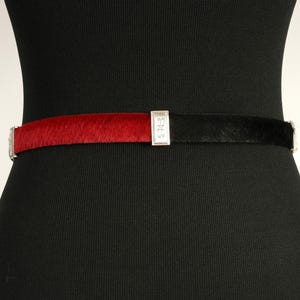 Puede incluir: Un cintur&oacute;n bicolor con una textura similar a la piel de color rojo y negro. El cintur&oacute;n est&aacute; dividido por una hebilla rectangular plateada con un dise&ntilde;o ornamentado. El cintur&oacute;n se abrocha con una hebilla plateada.