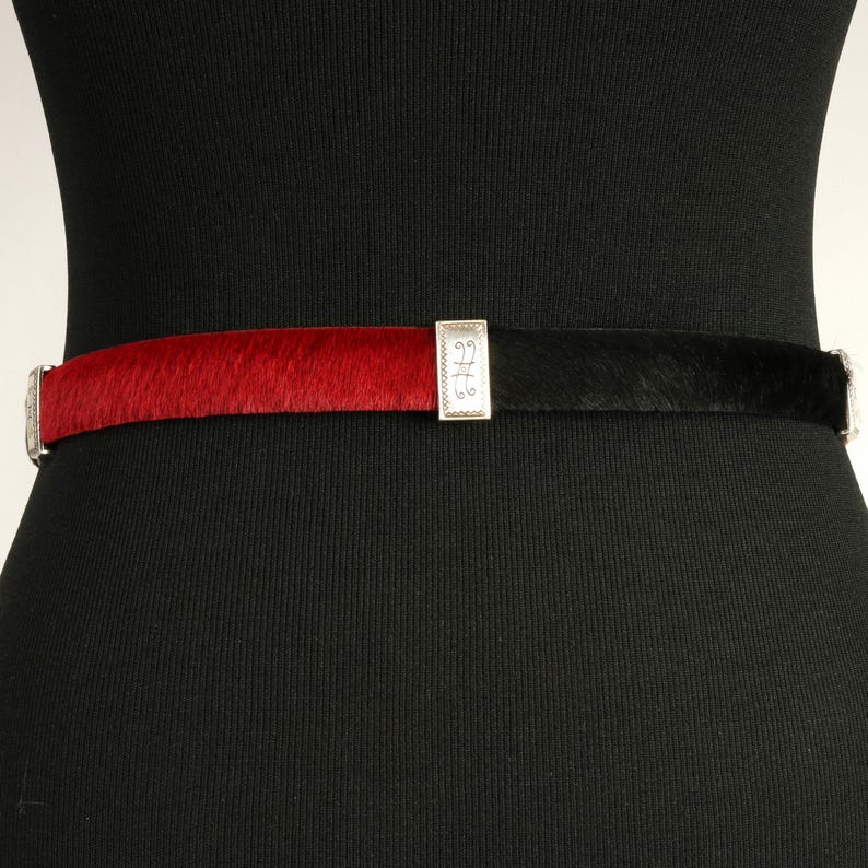 Puede incluir: Un cintur&oacute;n bicolor con una textura similar a la piel de color rojo y negro. El cintur&oacute;n est&aacute; dividido por una hebilla rectangular plateada con un dise&ntilde;o ornamentado. El cintur&oacute;n se abrocha con una hebilla plateada.