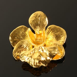 May include: A gold-colored, three-dimensional orchid flower with detailed petals. The flower is made of a shiny, reflective material, and the petals have a textured surface. The orchid is a decorative item.