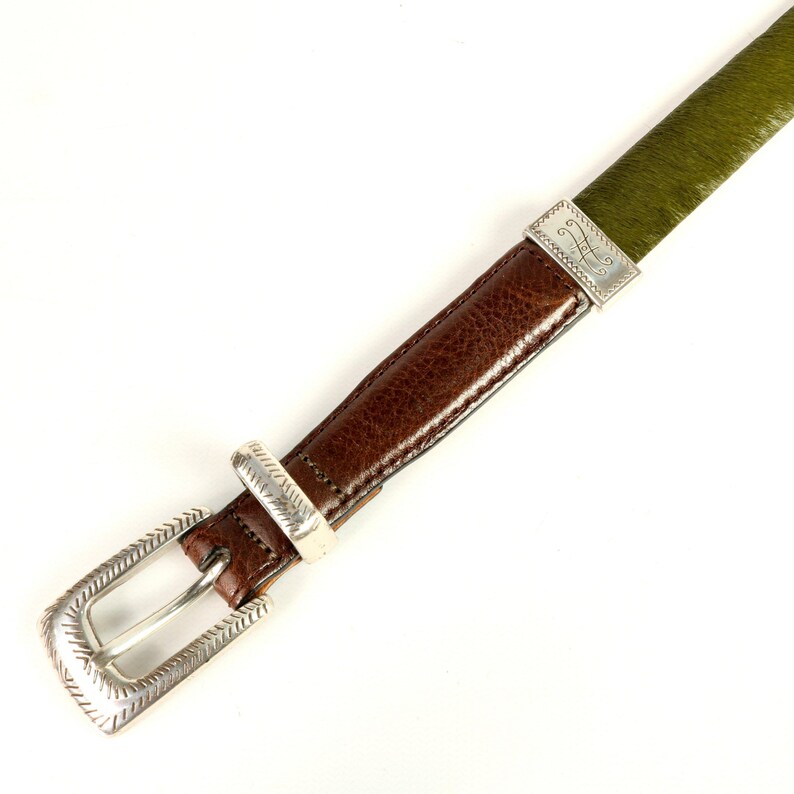 Puede incluir: Un cintur&oacute;n de cuero marr&oacute;n con hebilla y detalles plateados. El cintur&oacute;n presenta una secci&oacute;n verde texturizada y una correa de cuero marr&oacute;n. La hebilla y los detalles tienen un patr&oacute;n decorativo. El cintur&oacute;n est&aacute; inclinado sobre un fondo blanco.