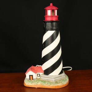 May include: A decorative lighthouse lamp with a black and white striped tower, a red top, and a small white house with a red roof. The lamp sits on a base with faux grass and a brown border.