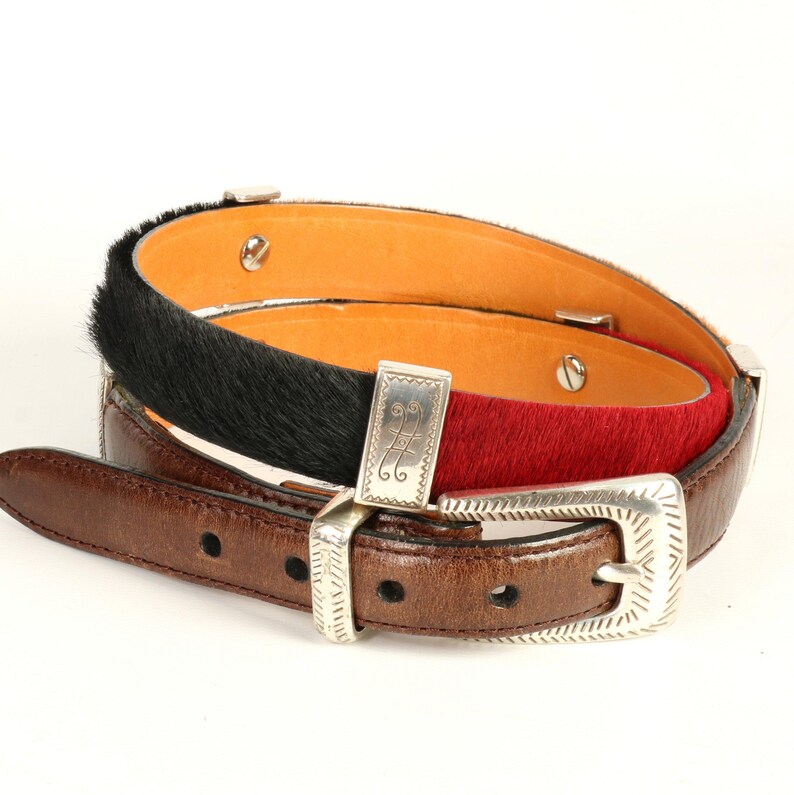 Puede incluir: Tres cinturones de cuero con hebillas plateadas. Uno es marr&oacute;n, otro tiene pelo negro y el tercero pelo rojo. Los cinturones est&aacute;n apilados, mostrando las diferentes texturas y colores.