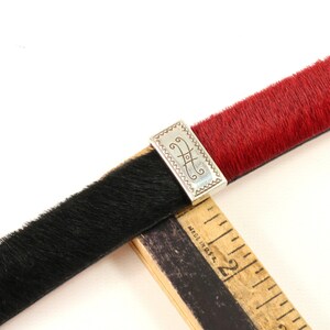 Puede incluir: Un cintur&oacute;n bicolor con una textura similar a la piel de color negro y rojo. El cintur&oacute;n est&aacute; conectado por una hebilla rectangular plateada con un dise&ntilde;o grabado. Una regla de madera se coloca sobre el cintur&oacute;n.