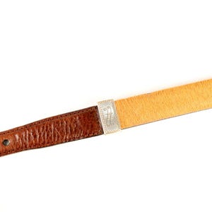 Puede incluir: Un cintur&oacute;n de cuero marr&oacute;n con una secci&oacute;n de pelo animal de color marr&oacute;n claro. El cintur&oacute;n presenta una hebilla plateada con un dise&ntilde;o ornamentado. La parte de cuero tiene un aspecto texturizado, sobre un fondo blanco.