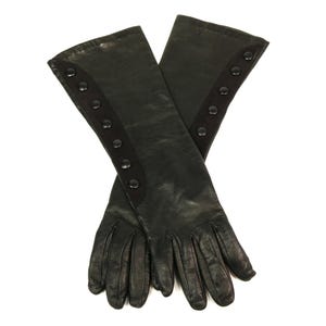 May include: A pair of long black leather gloves featuring a smooth finish and a vertical panel of five matching black buttons along the outer edge of each forearm. The gloves extend past the wrist and are positioned in a crossed arrangement.