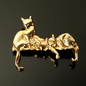 May include: A gold-toned metal brooch featuring two cats. One cat is in a seated upright position, while the second cat is lying down horizontally next to it. The surface of the metal has a polished, reflective finish with detailed contours.