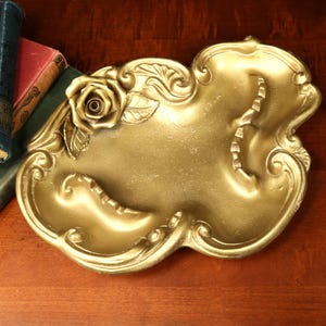 May include: A gold-colored, ornate decorative tray with a rose and leaf design. The tray has a unique, flowing shape with curved edges and a textured surface. The tray is likely made of metal and is suitable for display or serving.
