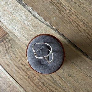 May include: A silver ring with an open, irregular, and fluid shape. The ring features a thin, polished metal band that curves into a continuous, non-symmetrical loop, resting on a circular gray velvet display stand with a central slit.