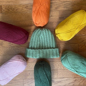May include: Six balls of yarn in various colors, including orange, yellow, burgundy, pink, and two shades of green, arranged around a green crocheted beanie.