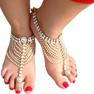 May include: Gold-toned foot jewelry with multiple strands of small, white beads and clear, sparkling crystals. The anklets feature a large, central crystal and a ring that connects to the toe. The jewelry is designed to be worn on the feet.