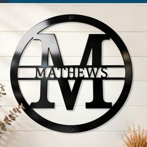 Personalized monogram door sign; circular black metal frame featuring M initial and MATTHEWS name for front door decor.