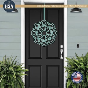 Turquoise metal mandala door wreath on a black front door, flanked by greenery—geometric outdoor front door decor.
