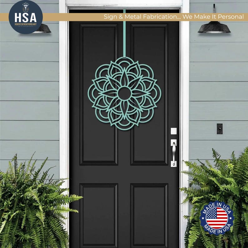 Turquoise metal mandala door wreath on a black front door, flanked by greenery—geometric outdoor front door decor.