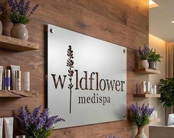 May include: A rectangular silver sign mounted on a wood-paneled wall features the text wildflower medispa in black lettering with a stylized floral graphic. The sign is surrounded by multiple shelves holding white bottles and potted lavender plants.