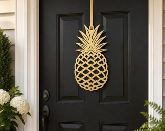 May include: A gold-colored, laser-cut metal pineapple wall hanging suspended by a matching gold strap. The pineapple features a geometric, open-work lattice pattern on the body and stylized, pointed leaves at the top, centered on a black door.