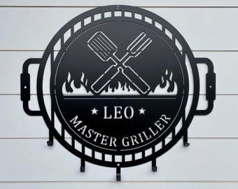 May include: A circular black metal wall sign shaped like a grill, featuring a cutout design of crossed spatula and fork utensils above flames. The text LEO and MASTER GRILLER is centered, with four small hooks attached to the bottom edge for hanging.