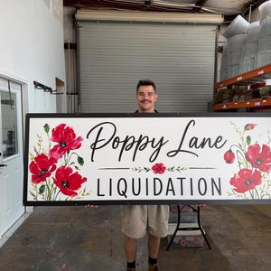 May include: A large rectangular sign with a white background and a black frame. The text "Poppy Lane" is written in a black script font above the word "LIQUIDATION" in a black sans-serif font. Red poppy flowers are painted on both sides.
