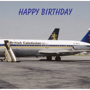Puede incluir: Un avión blanco con detalles azules y amarillos y las palabras "British Caledonian" en el lateral. El avión está en una pista de aterrizaje con un cielo azul de fondo. Las palabras "Happy Birthday" están escritas en letras azules en la parte superior de la imagen.